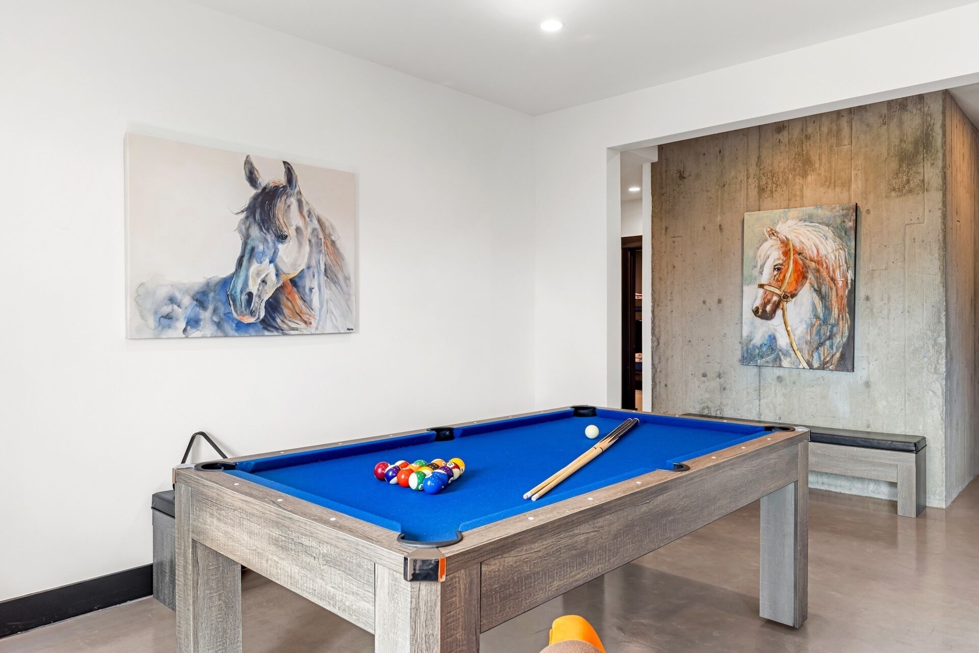Une table de billard avec des billes et des queues, entourée de murs blancs ornés de deux tableaux représentant des chevaux.