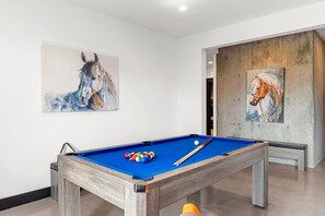 A pool table with balls and cues, surrounded by white walls with two horse paintings.
