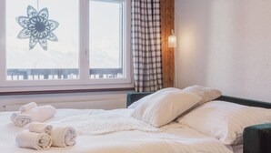Un letto rifatto con cura, asciugamani arrotolati, una finestra con un oggetto decorativo e una tenda a quadri.