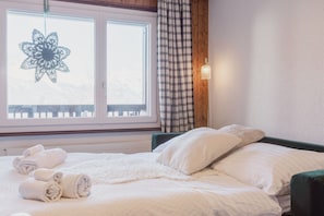 A neatly made bed with rolled towels, a window with a decorative item, and a checkered curtain.