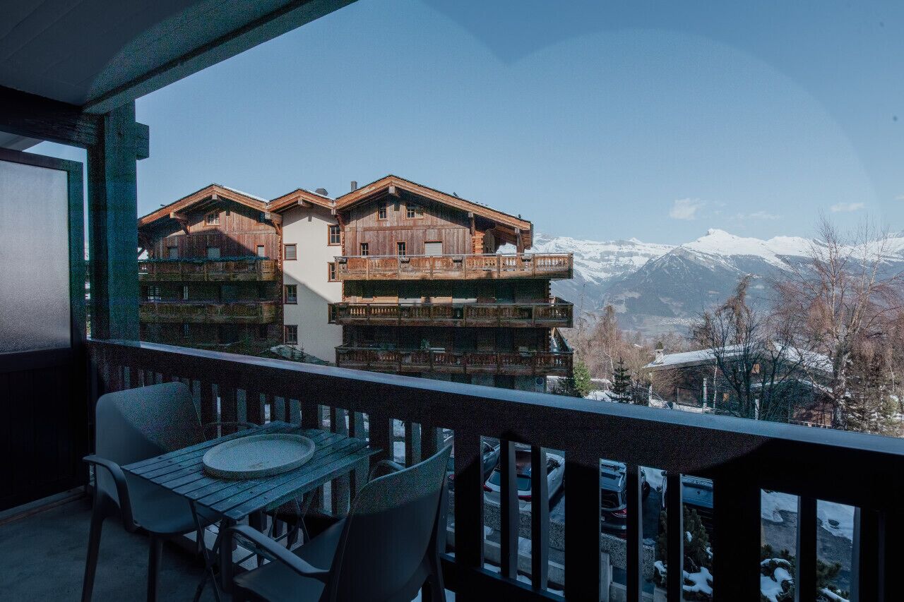 A balcony with a table and chairs, overlooking a mountain landscape with snow-covered peaks.