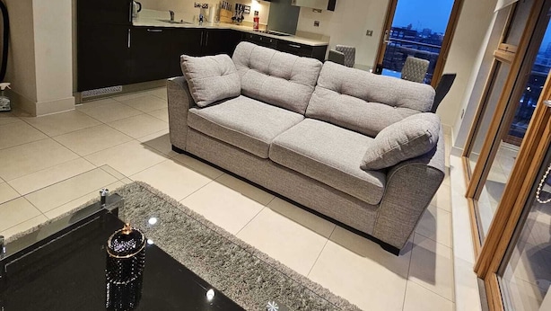 Un salon moderne avec un canapé gris, une table basse noire et une cuisine en arrière-plan.