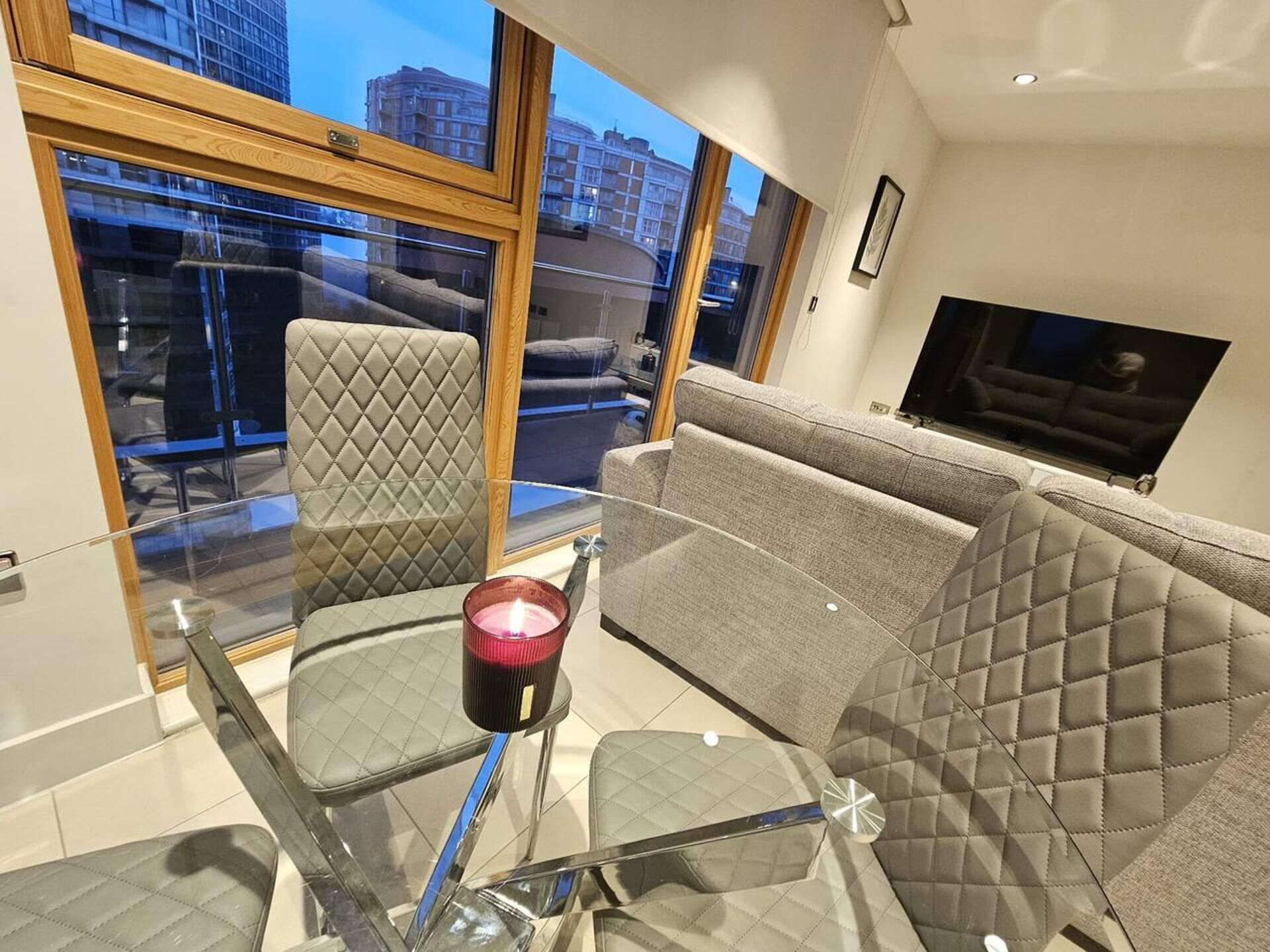 A modern living room with a glass table, cushioned chairs, a grey sofa, and a TV on the wall.