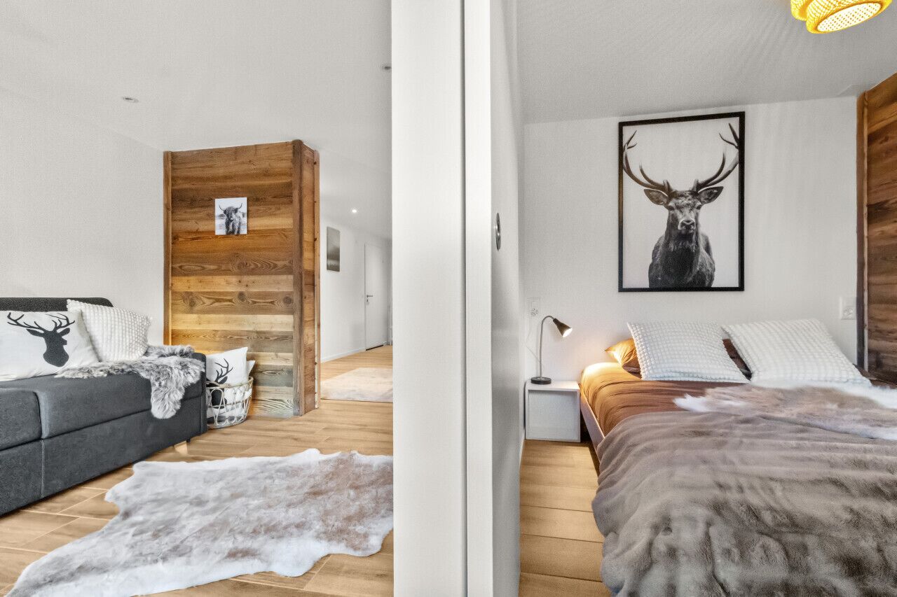 A modern bedroom with a wooden accent wall, a large framed deer picture, a grey sofa with deer-themed pillows, a fur rug, and a wooden floor.