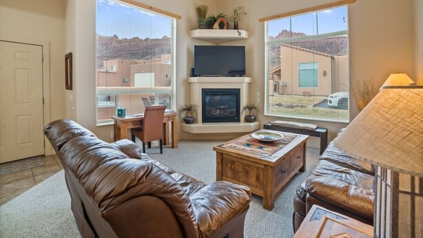 A living room with a fireplace, leather sofas, a coffee table, and a view of a mountainous landscape. - Rim Village X3 - Large Patio & Private Hot Tub (Moab)