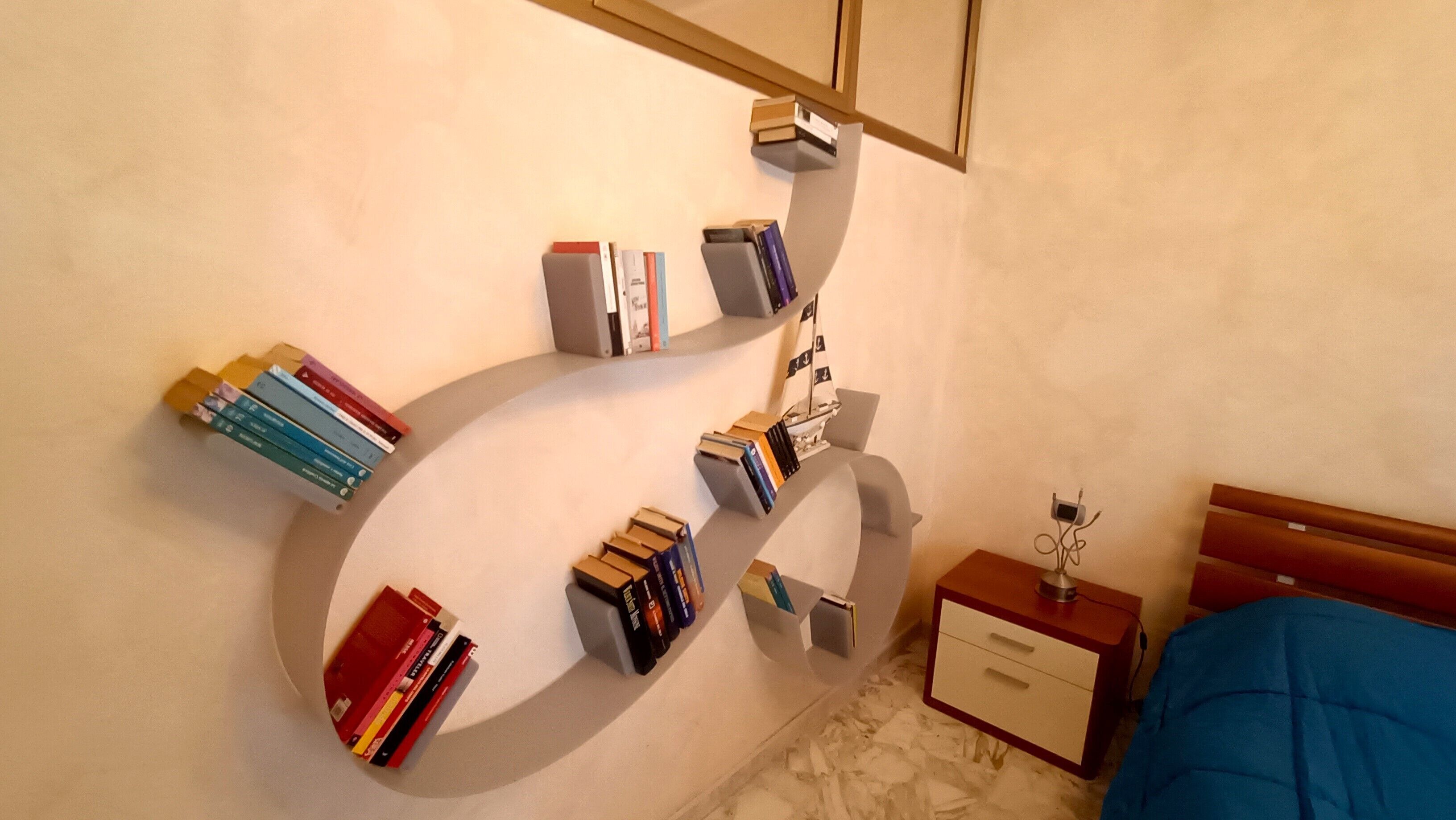Ein Schlafzimmer mit einem gebogenen Bücherregal, einem hölzernen Nachttisch und einem Bett mit blauer Decke.