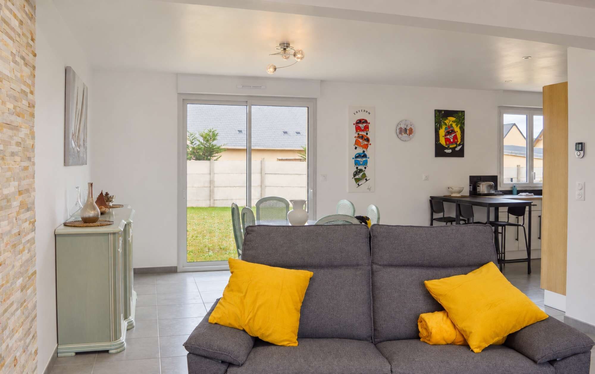 A modern living room with a grey sofa and yellow cushions, a dining area with a table and chairs, and a kitchen visible through an open door.