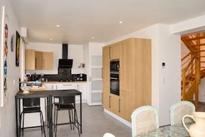 A modern kitchen with wooden cabinets, a black countertop, and a dining area with a glass table and wicker chairs. - Les Embruns, Maison Neuve Près des Plages (Asnelles)
