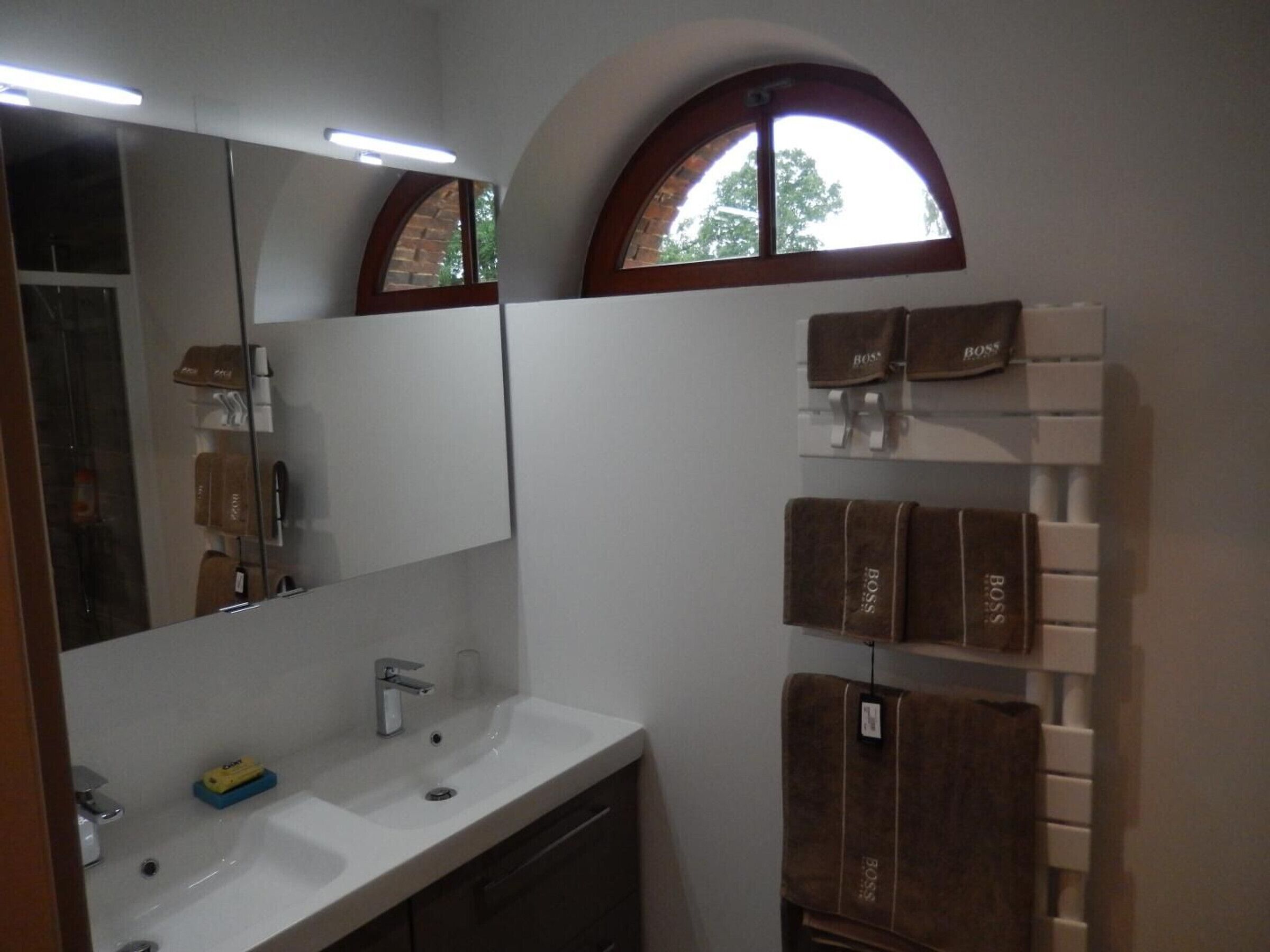 Une salle de bain avec un lavabo blanc, un miroir et une fenêtre surmontée d’une arche en bois. Des serviettes sont suspendues sur un porte-serviettes.