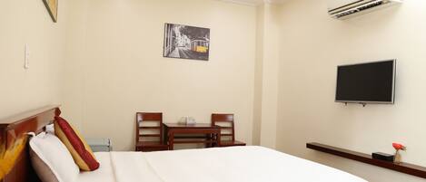 A hotel room with a bed, a desk with chairs, a flat-screen TV, and an air conditioning unit.