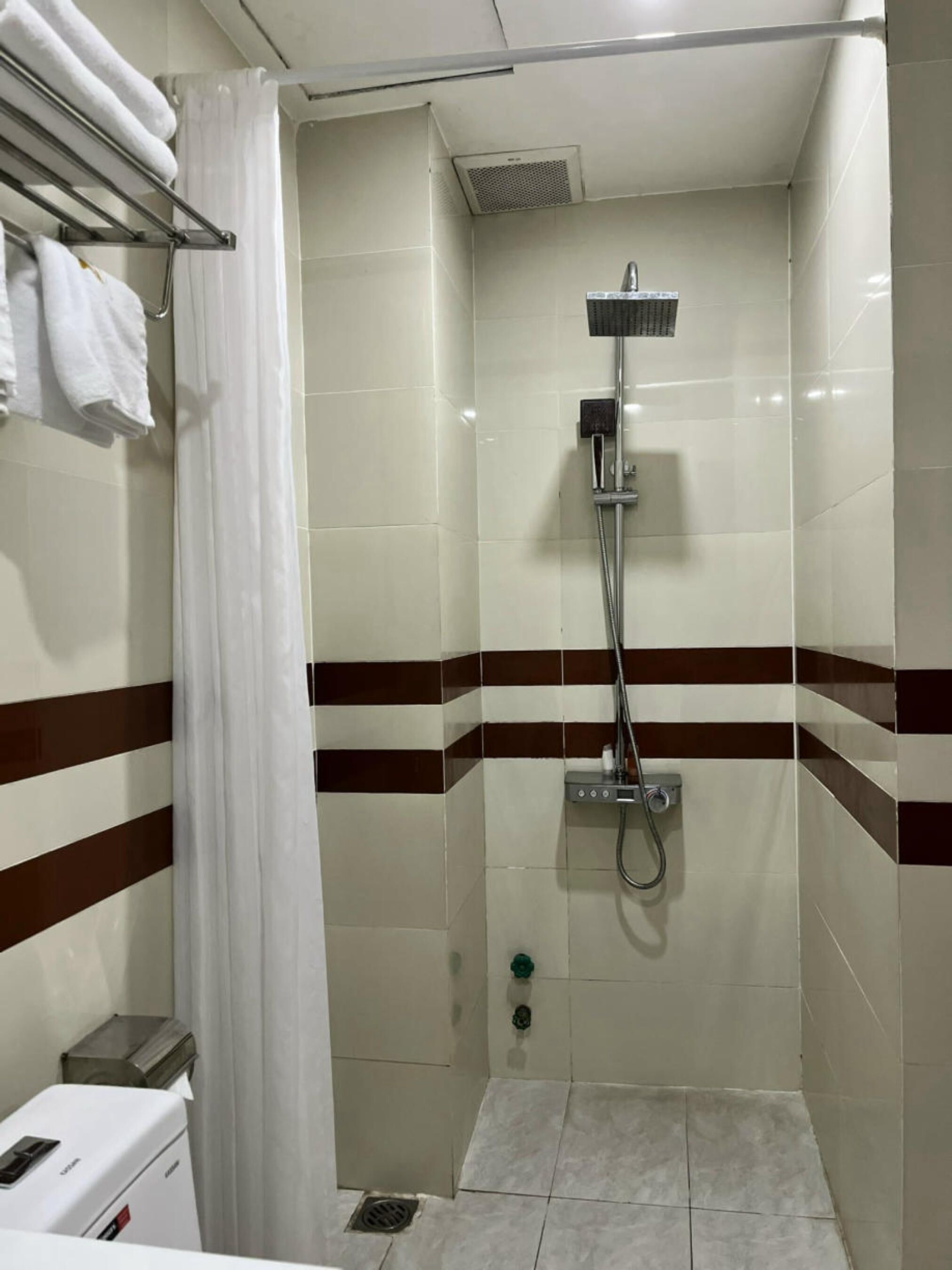 A small, tiled shower room with a showerhead, a towel rack, and a trash bin.