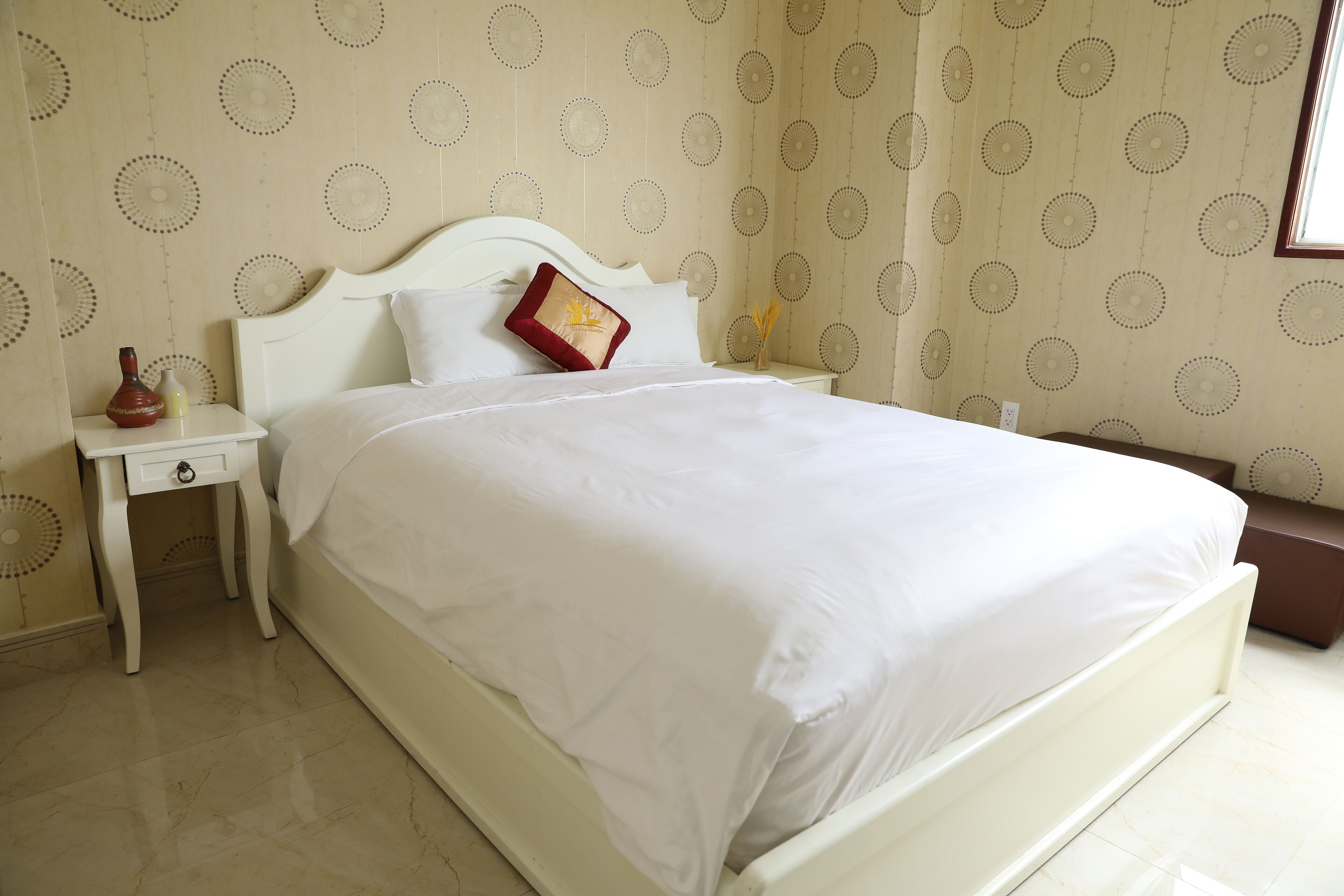A neatly made bed with white bedding and a red accent pillow, a white nightstand with a vase, and a patterned wallpaper.