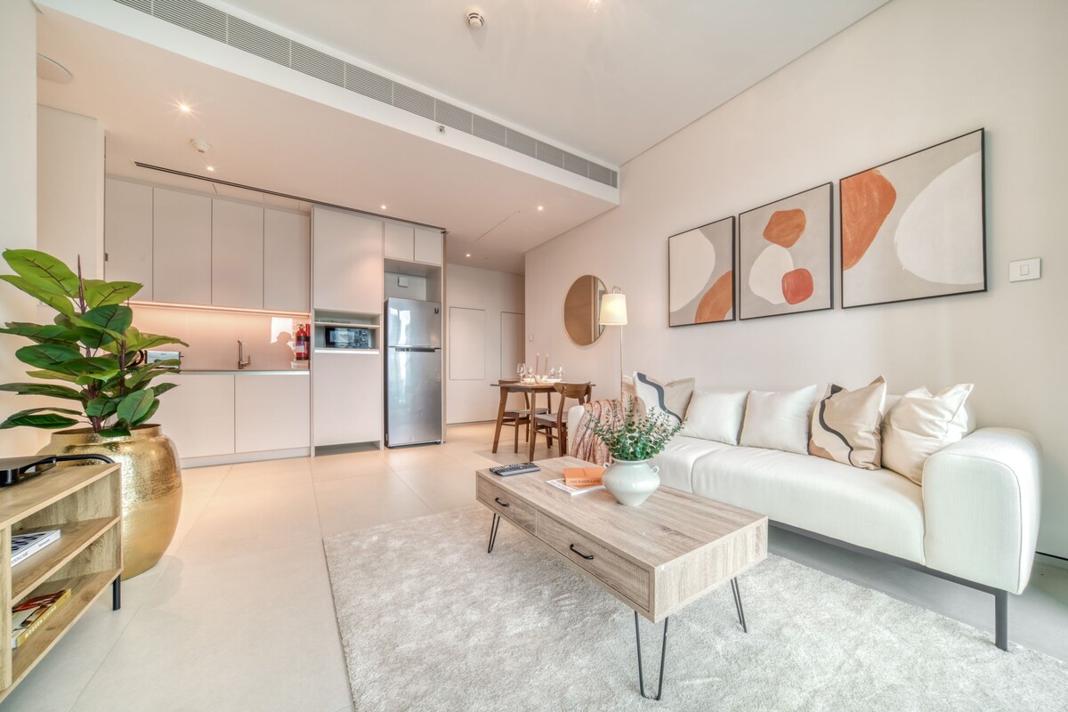A modern living room with a white sofa, a coffee table, and a dining area with a table and chairs. There is a kitchen visible in the background.