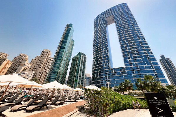 A beachside resort with sun loungers, palm trees, and modern high-rise buildings in the background. - Address JBR - Mint Stay (Dubai)