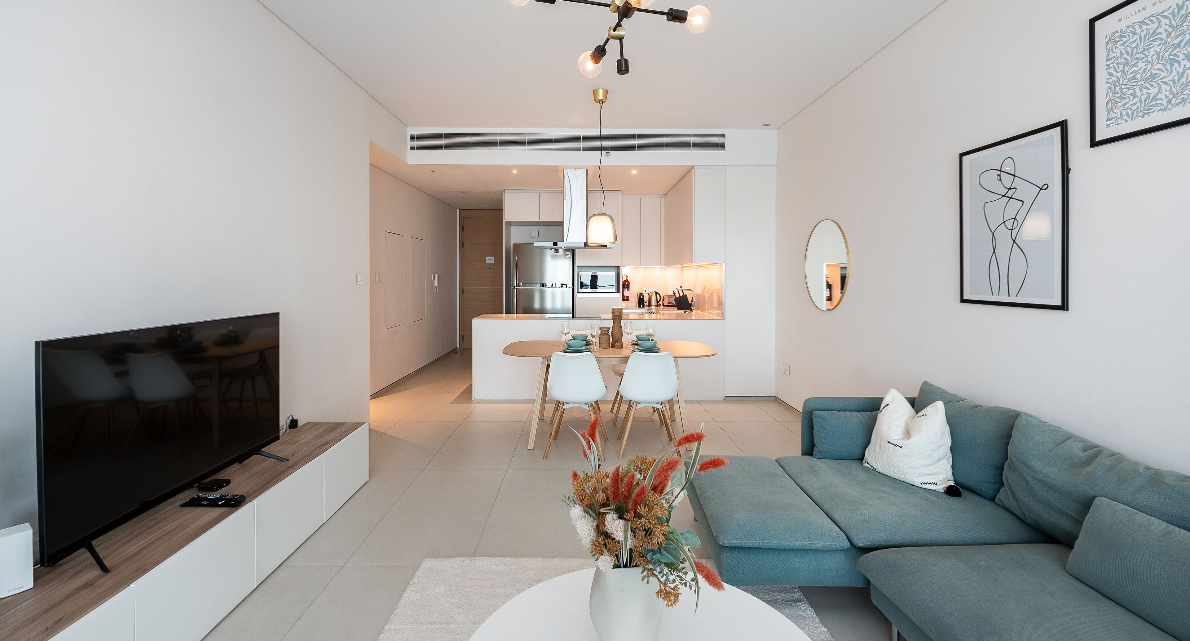 A modern living room with a flat-screen TV, a teal sofa, and a dining area with a kitchen in the background.
