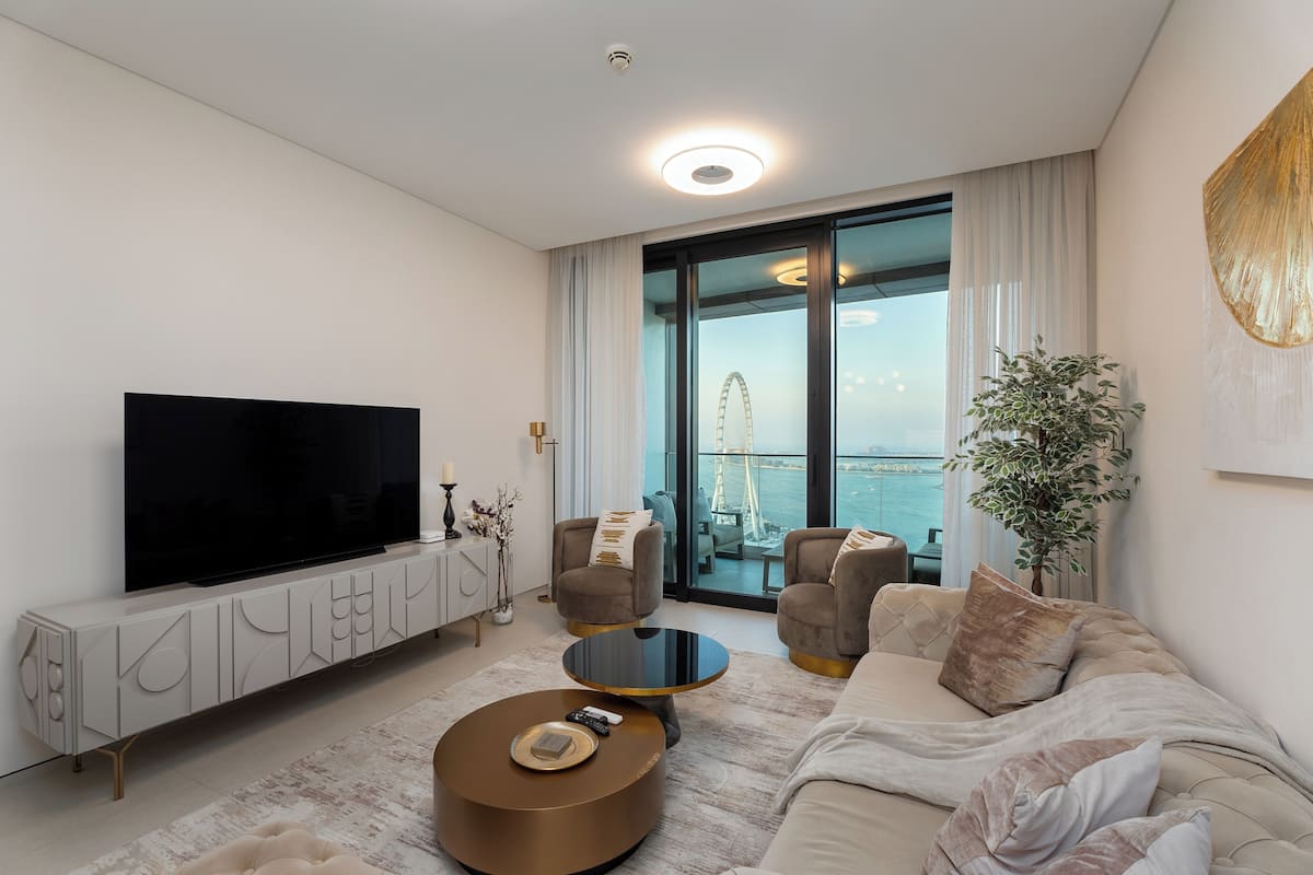 A modern living room with a large flat-screen TV, comfortable seating, a coffee table, and a view of a Ferris wheel through a glass door.