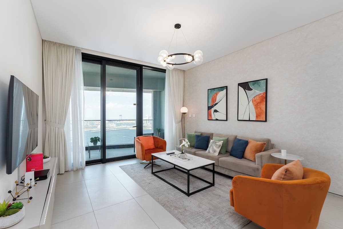 A modern living room with a flat-screen TV, a coffee table, orange armchairs, a sofa, and a large window offering a view of the city.