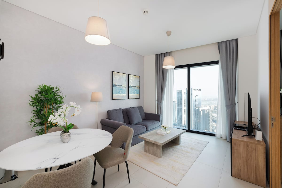 A modern living room with a sofa, a dining table with chairs, a coffee table, a TV on a wooden stand, and a large window with a city view.
