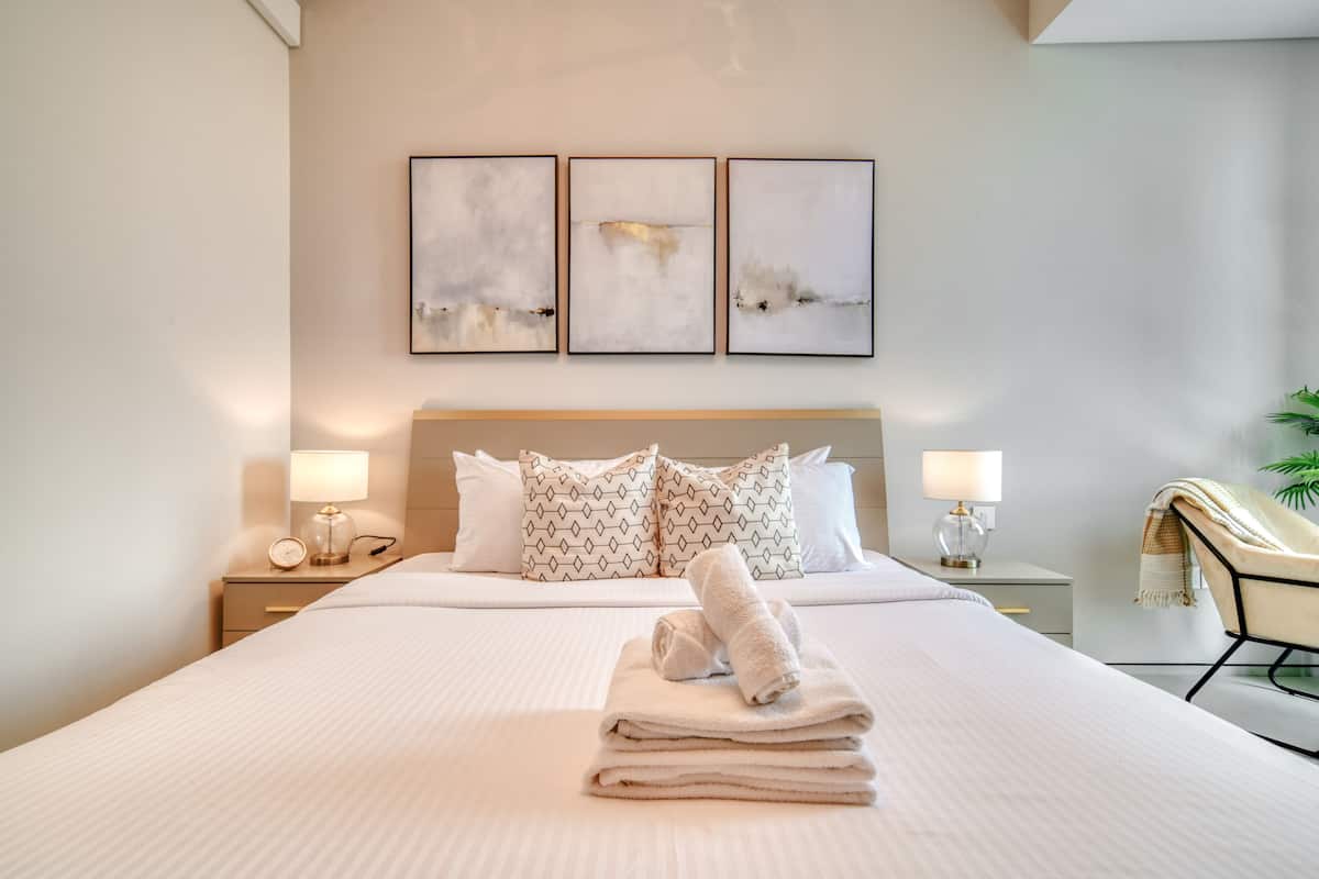 A neatly made bed with a stack of towels, a bedside table with a lamp, and a chair with a blanket.