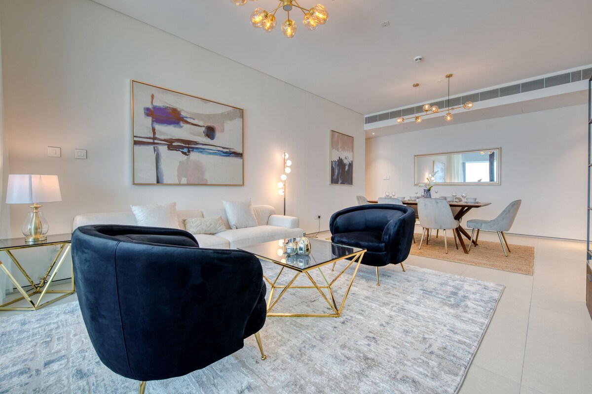 A modern living room with a white sofa, two dark armchairs, a glass coffee table, and a dining area with a table and chairs.