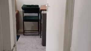 A narrow hallway with a door on the left, a green chair and a small table in the background, and a doormat with a pair of flip-flops on the floor. - Amazon one bedroom apartment  (Nakuru)