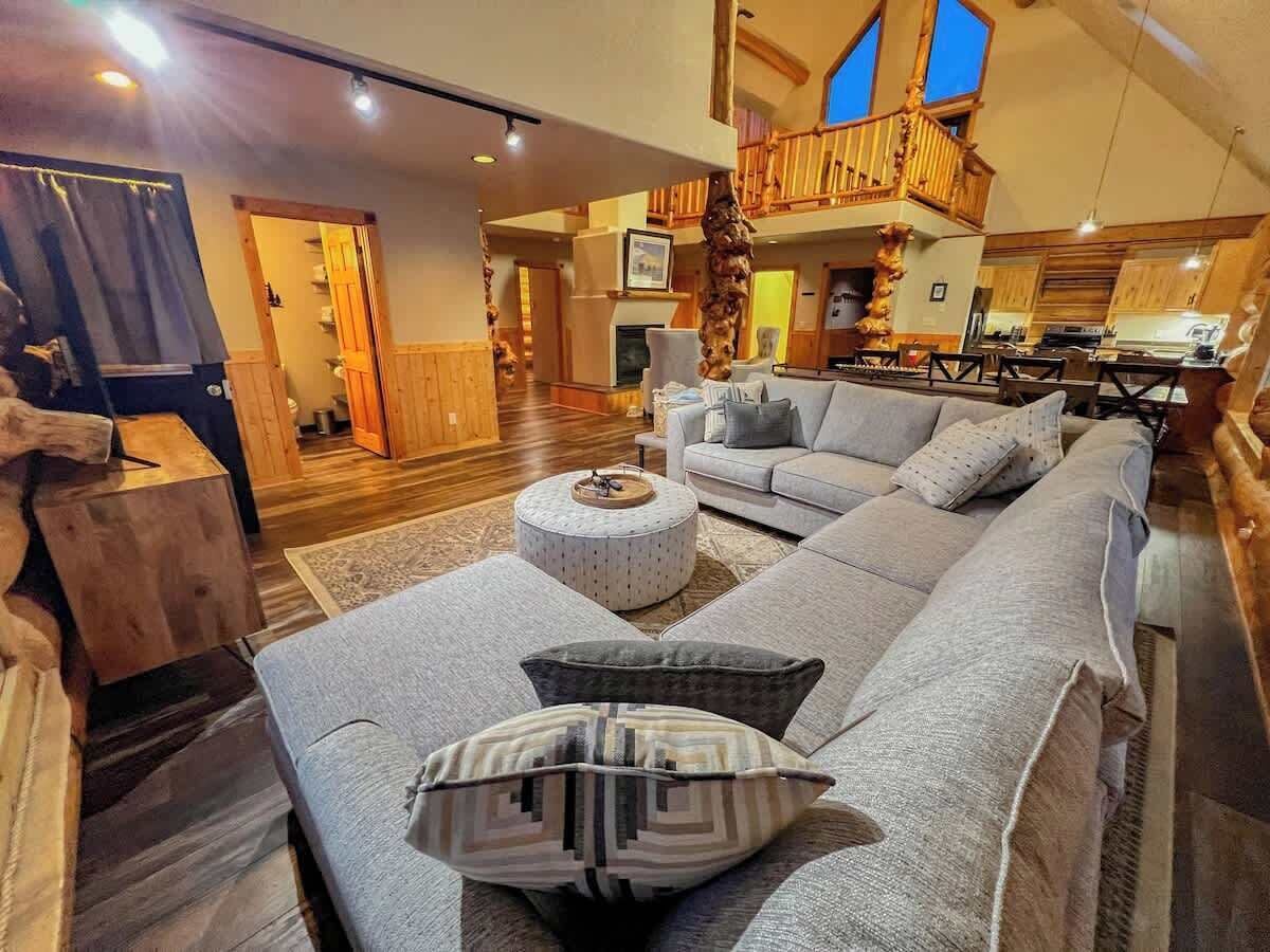Amplia sala de estar con un sofá en forma de L, un otoman redondo y una mesa de centro de madera. Se observa un balcón con barandillas de madera y vista al cielo al fondo.