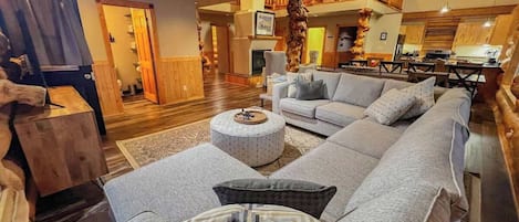 Amplia sala de estar con un sofá en forma de L, un otoman redondo y una mesa de centro de madera. Se observa un balcón con barandillas de madera y vista al cielo al fondo.