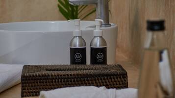 Une salle de bain avec une baignoire, deux flacons de savon liquide, une serviette, un tapis tissé et une plante.