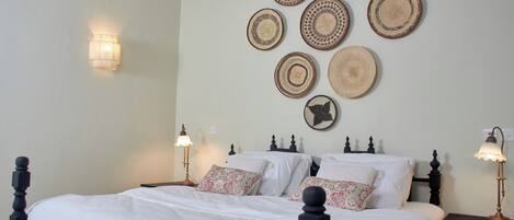A neatly made bed with white linens and patterned pillows, a wall-mounted lamp, and a series of woven wall hangings.
