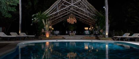 A nighttime pool area with a thatched-roof structure, lounge chairs, and a clear pool.