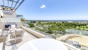A balcony with outdoor seating, clear glass railings, and a view of a cityscape and greenery.