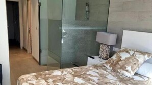A modern bedroom with a glass-enclosed shower, a bed with a floral bedspread, and a nightstand with a lamp.