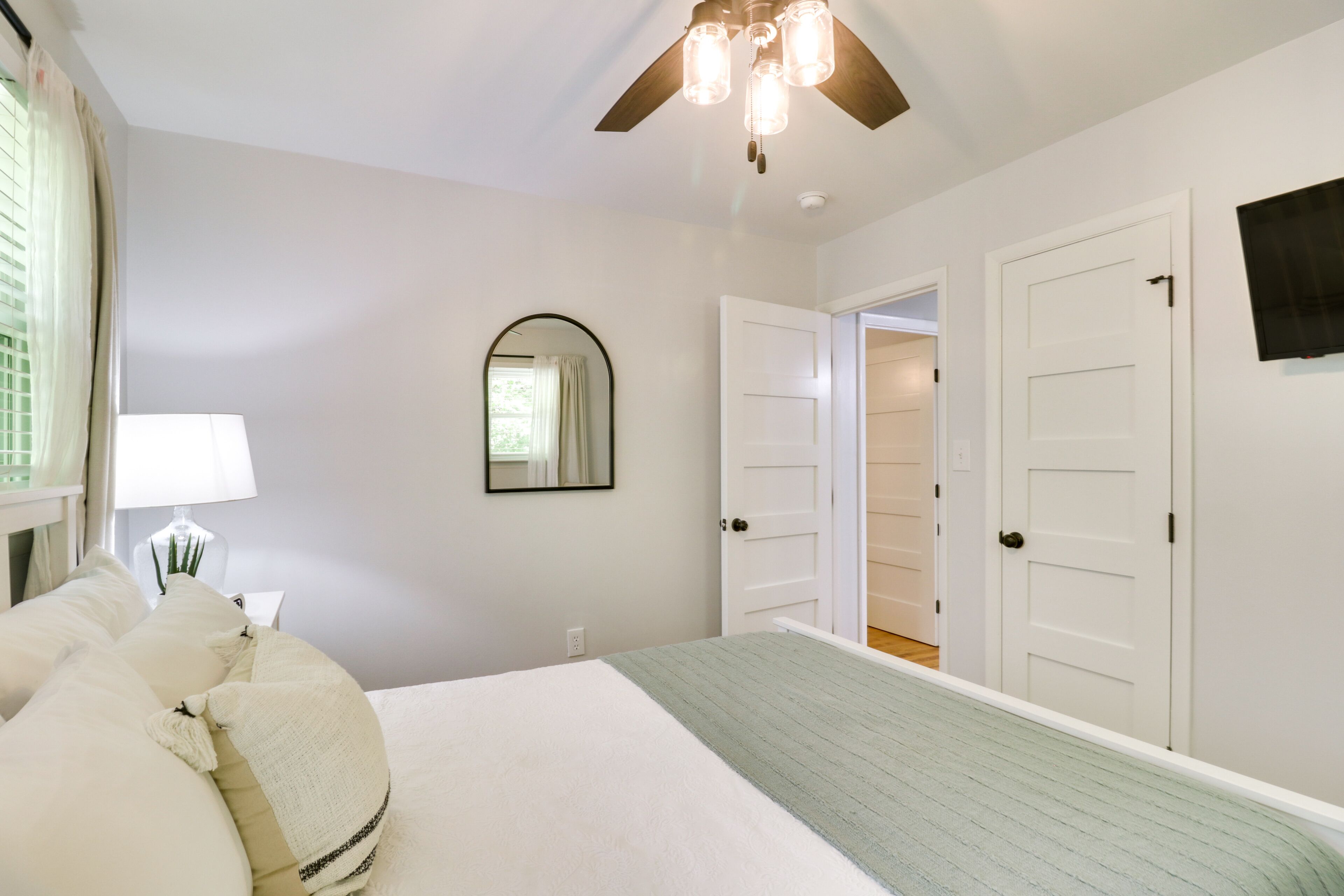 Un dormitorio con cama, ventilador de techo, espejo y puerta que da a otra habitación.