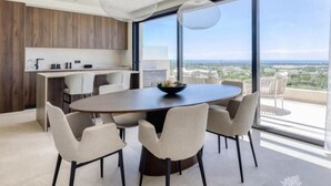 A modern dining area with a round table and chairs, a kitchen with wooden cabinets, and large windows offering a city view. - Penthouse Limonero 30 (Dehesa de Campoamor)