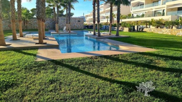 A swimming pool surrounded by palm trees and a well-maintained lawn, with a stone wall and a modern building in the background. - Penthouse Limonero 30 (Dehesa de Campoamor)
