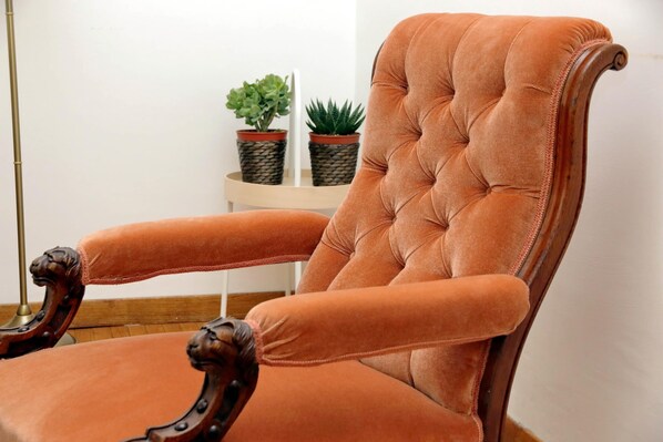 A tufted armchair with wooden armrests and a cushioned seat, positioned next to a wooden side table with two potted plants.