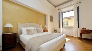 A bedroom with a large bed, wooden flooring, a nightstand with a lamp, a small table with a plant, and a view through an open door. - Treasurerome Spanish Steps Family 4BR (Rome)