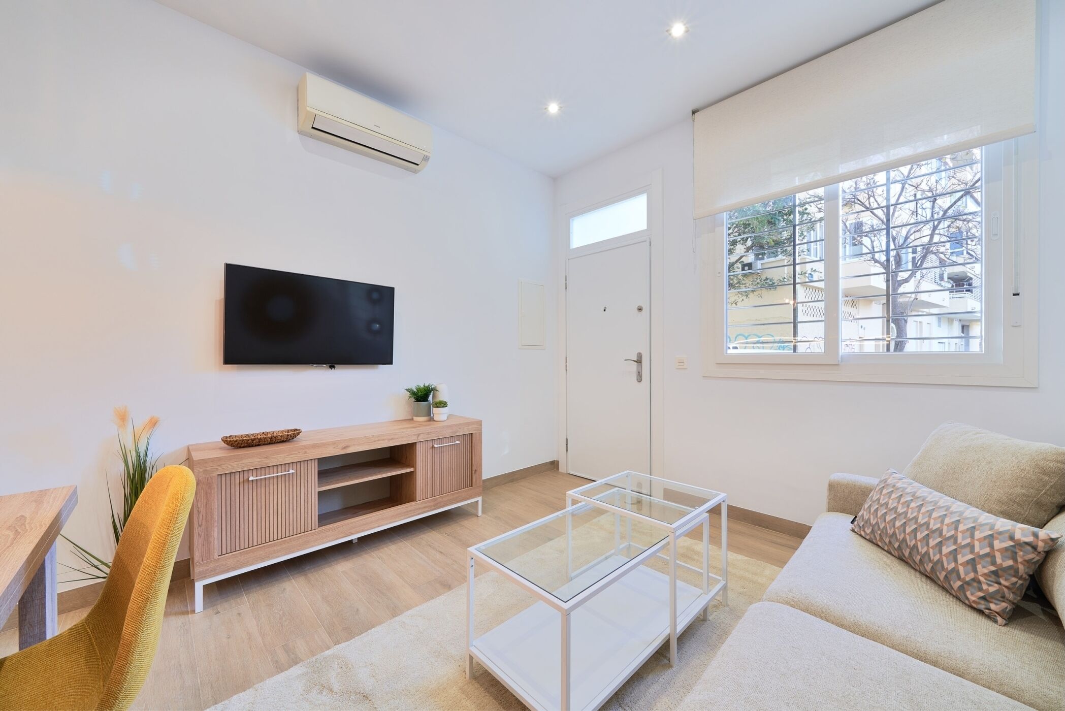 A modern living room with a flat-screen TV, wooden entertainment unit, glass coffee table, and a sofa with patterned cushions.
