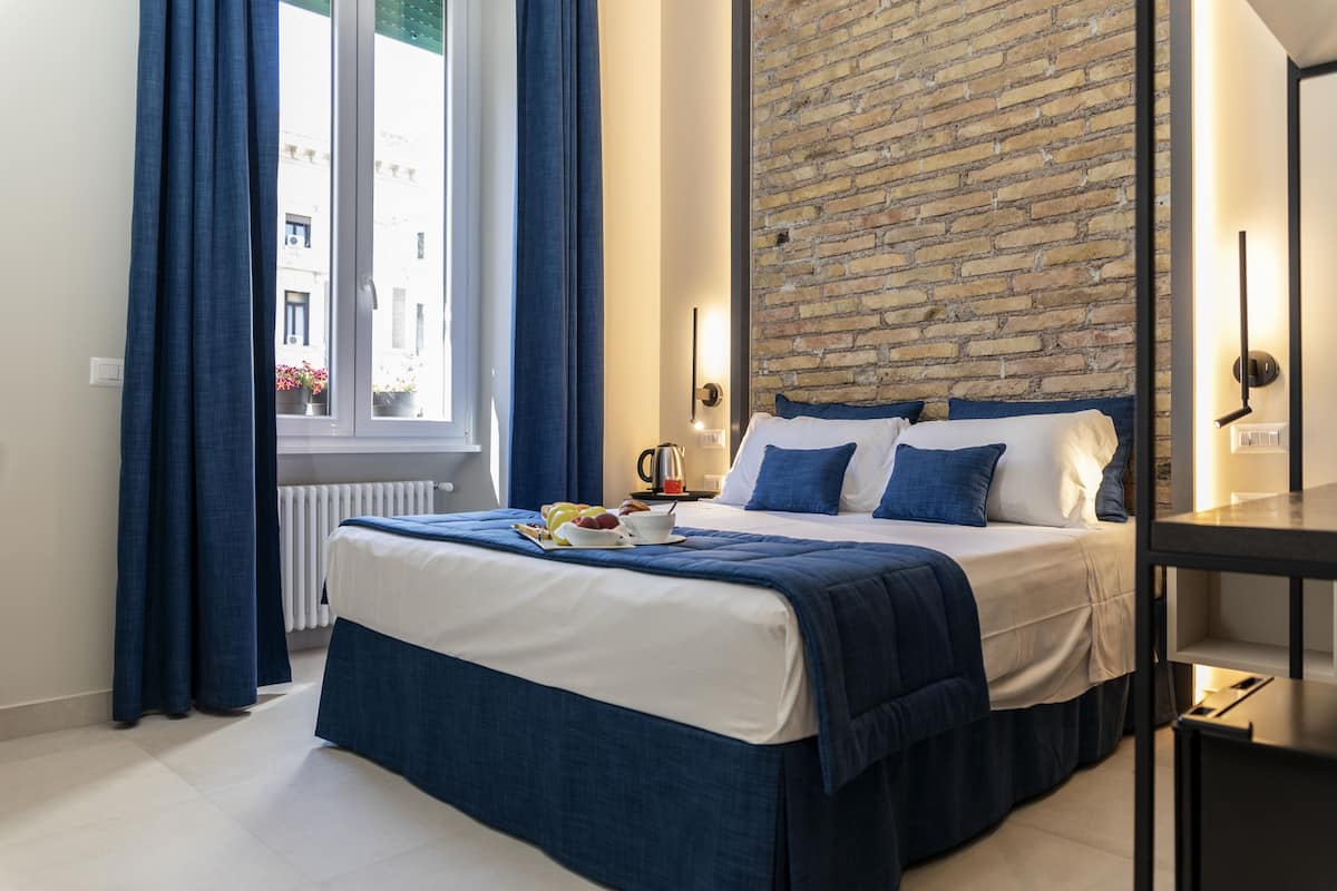 A modern hotel room with a large bed, blue curtains, a brick accent wall, and a view of buildings outside.