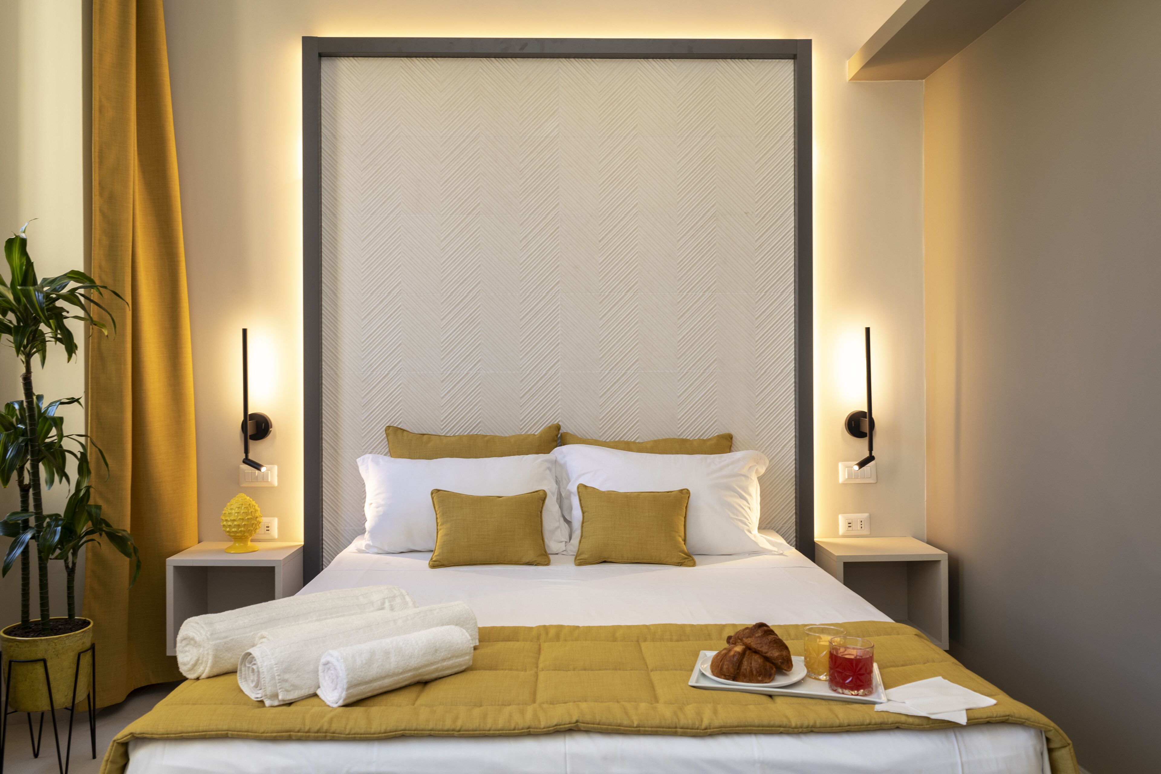 Een netjes opgemaakt bed met wit en mosterdgeel beddengoed, een nachtkastje met een lamp en een potplant.