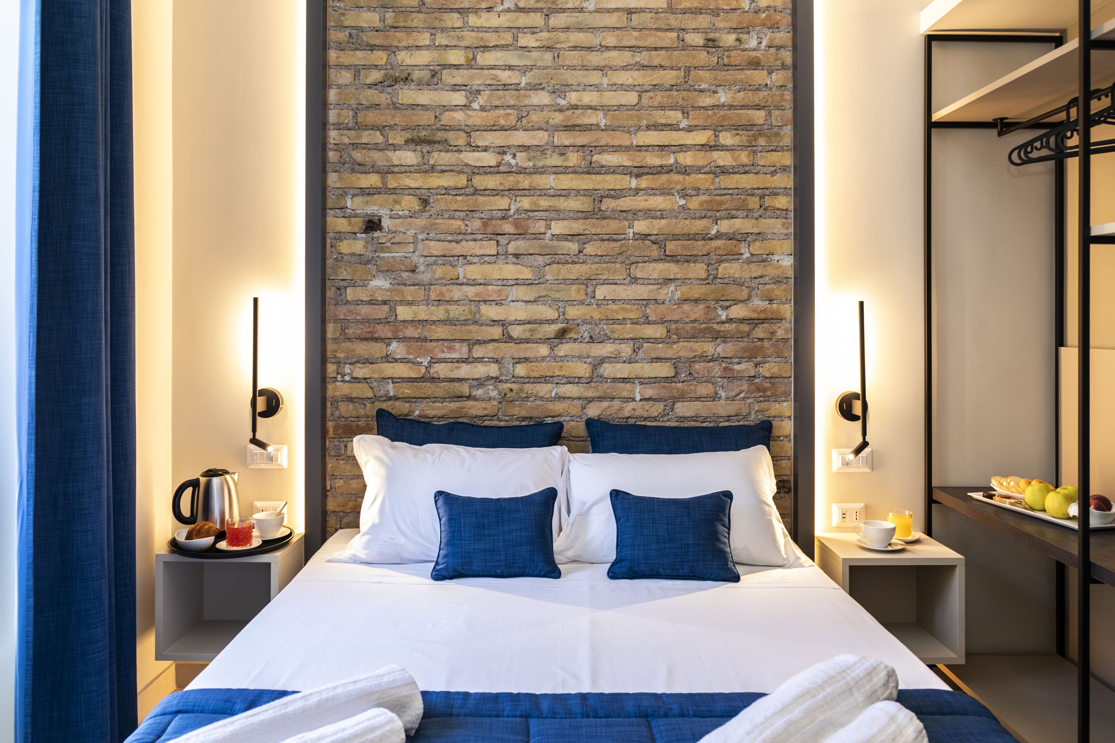 A hotel room with a brick wall, a bed with white and blue bedding, two bedside tables with lamps, and a small shelf with fruits and a cup.