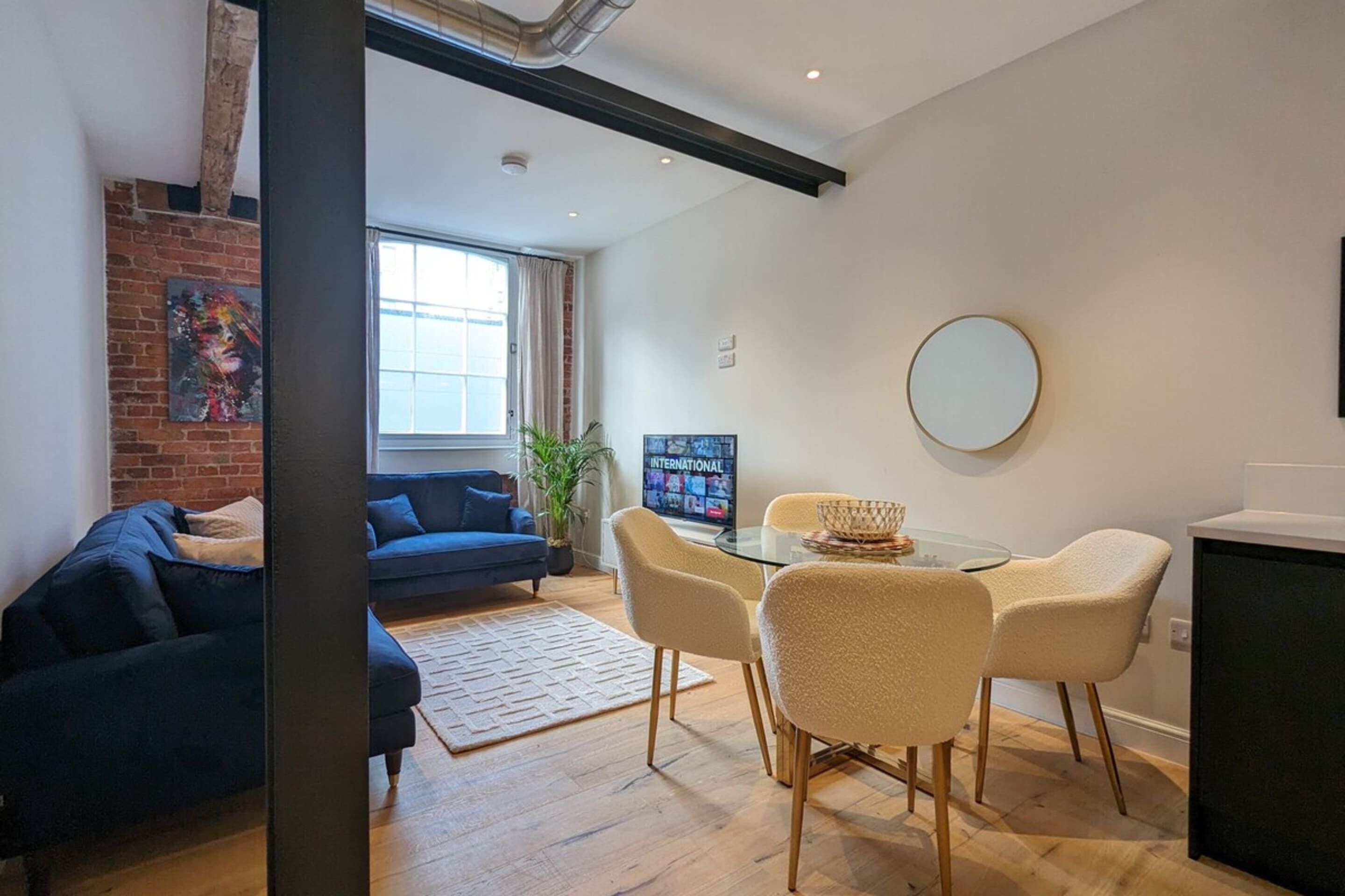 A modern living room with a blue sofa, a glass dining table with chairs, a TV, and a brick wall with artwork.