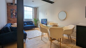Ein modernes Wohnzimmer mit einem blauen Sofa, einem Glastisch mit Stühlen, einem Fernseher und einer Ziegelwand mit Kunstwerken.