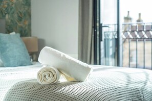 A neatly made bed with a rolled white towel on it, a window with a view of buildings, and a blue pillow. - Superb 3-bed house with parking in Bath (Bath)