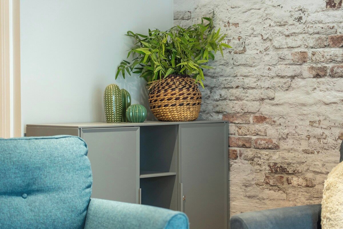A room with a grey sofa, a blue armchair, a wicker plant pot, and a brick wall.