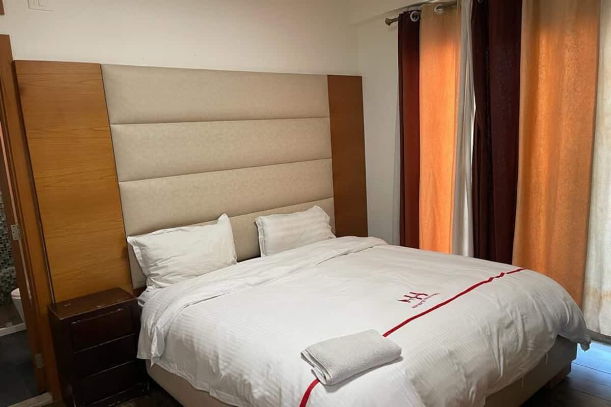 A neatly made bed with white linens and a red accent, a wooden headboard, and a nightstand with a lamp.