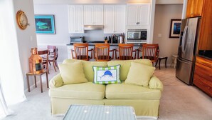 A living room with a green sofa, a white coffee table, and wooden chairs. There is a kitchen in the background with white cabinets and a refrigerator. - BAY FRONT HOME WITH AMAZING VIEWS.  MINUTES TO THE BEACH AND STEPS TO THE POOL  (Fenwick Island)