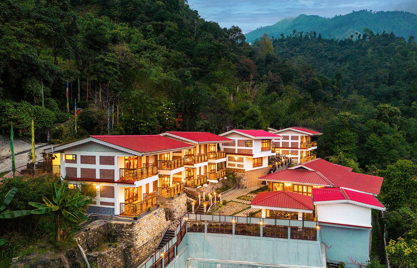 A hotel with multiple buildings, each with red roofs and white walls, situated on a hillside with lush greenery and mountains in the background.