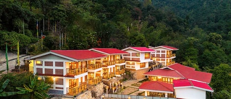 A hotel with multiple buildings, each with red roofs and white walls, situated on a hillside with lush greenery and mountains in the background.