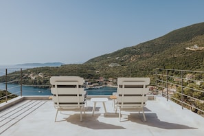 Une terrasse avec deux chaises blanches et une petite table, donnant sur une zone côtière avec des bâtiments et de la végétation.